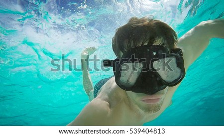 Handsome man is taking self picture while driving and exploring underwater life in the deep ocean. He's wearing black goggles and swimwear. Around him are beautiful fishes and underwater scenes.