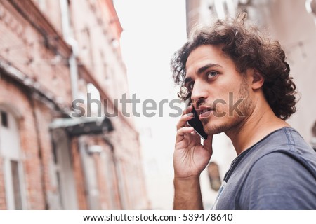 Handsome man with curly hair wearing t-shirt talks on the phone and smiling cheerfully while walking around in old city streets. Connectivity and technologies concept. Copy space