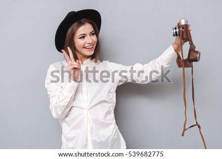 Happy young girl in hat making selfie with retro camera and showing peace sign isolated on a gray background