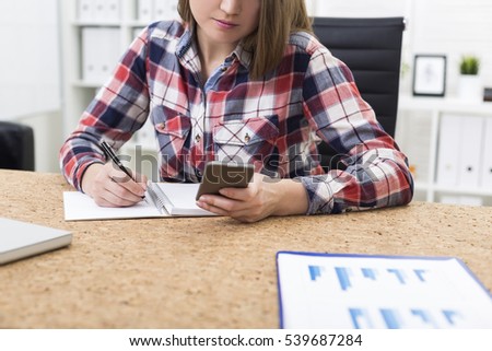 Close up of a girl with long brown hair in a red and black shirt looking at her smart phone screen and making notes in office