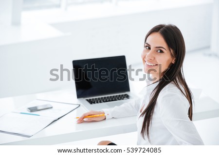 Back view of smiling business woman sitting by the table with laptop and looking at camera in office