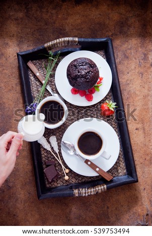 Black coffee in white cups and chocolate muffin with red berries on a dark wooden tray, hand pouring cream