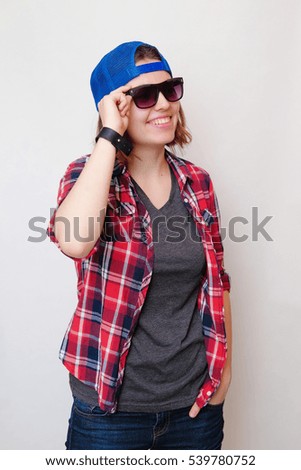 Hipster girl with short hair in a red plaid shirt, gray t-shirt, blue jeans, dark sunglasses, a blue cap, a black bracelet on a white background smiling and holding his hand over his glasses. 