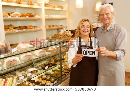 Stop by for homemade delicious bread! Loving senior couple embracing holding a sign together on the opening day of their small business senior couple owners of the bakery smiling to the camera