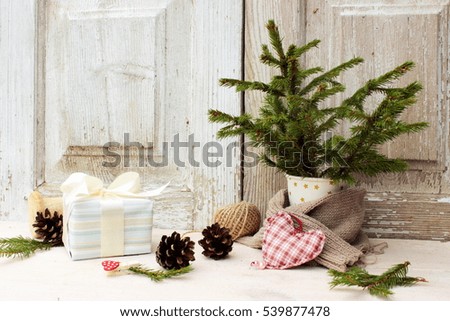 Pretty christmas composition with small natural christmas tree in a paper cup, cute gift box wrapped with blue paper with satin ribbon, some piny cones on white wooden background. Holiday decor
