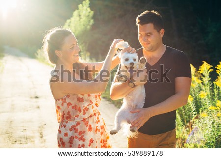 Young happy couple in love with dog having fun outdoors in lupines at sunset. Pretty girl in dress with young man in mustard shorts and black shirt playing with maltese. Beautiful light. 