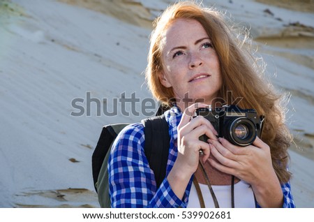 Young beautiful woman backpacker with red hair and freckles traveling in the desert and making pictures with vintage film camera. Sandy dunes background. Travel, adventure, freedom concept. 