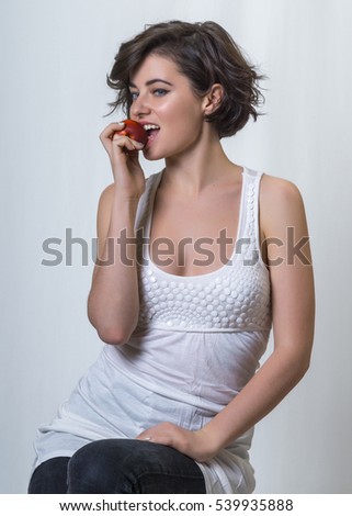 Young pretty brunette girl in white top takes a bite of the red apple on white background 