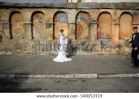 Beautiful bride in elegant dress stands before old orange wall and looks at groom