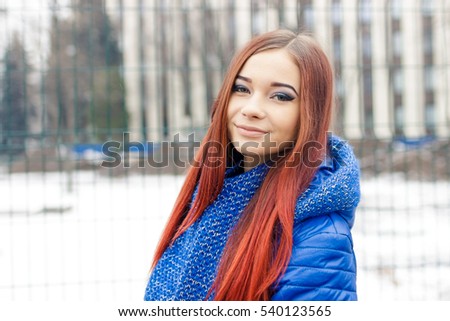 Pretty white brunette girl blow snow flakes from her frozen hands in snowy winter park.Good cold weather.Attractive young female wear knit earmuffs on her head,warm red scarf.Beautiful girl in blue.
