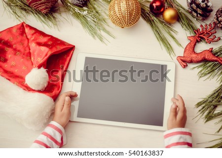 Happy Christmas background. Top view on child hands using tablet computer with toys and Santa Claus hat on light wooden table. Modern festive decoration ready for joyful message or shopping time
