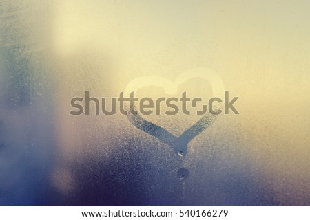 Abstract blurred love heart symbol drawn by hand on the wet frozen door window glass with sunlight background. Closeup of emotional image