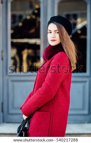 Stylish fashion brunette girl in red coat and black beret, posing over old gray door. Beauty girl in stylish coat. Model looking at camera. Fashion look.