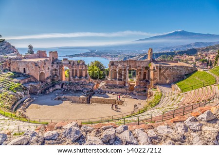 Ruins of the Ancient Greek Theater in Taormina, Sicily with the double smoke tail of the Etna extending over the the Giardini-Naxos bay of the Ionian Sea in the morning sun shine. Royalty-Free Stock Photo #540227212