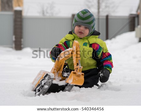 Cute cheerful child dressed in winter overalls playing excavator on white snow