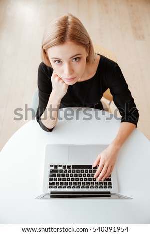 Top view photo of young cheerful woman worker sitting in office while using laptop computer and typing by keyboard. Looking at camera.