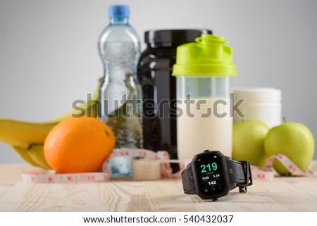 A set for a sportsman to be in shape. Sports watch for being organized, fruits as oranges, apples and bananas, shaker with protein cocktail, cans with sport supplements and mineral water on the desk.