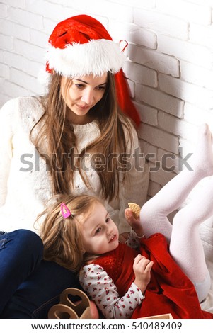 young cute little blonde girl in christmas red dress lying on her mothers lap in xmas hat and long brunette hair on white brick wall background