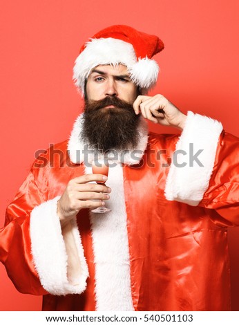 handsome bearded santa claus man with long beard on surprised face holding glass of alcoholic shot in christmas or xmas sweater and new year hat and touching mustache on red studio background