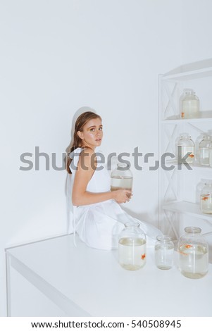 Cute young woman sitting on white table and holding gold fish in jar in the room