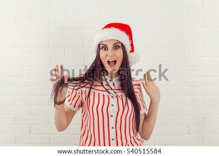 Surprised excited happy woman wearing red santa claus hat looking shocked by what she saw isolated white bricks background. Positive emotion