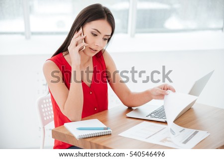 Woman talking at phone and sitting by the table with laptop in office