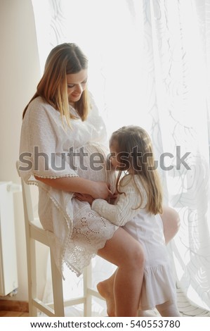 Charming little girl hugging her pregnant mother. The woman in a beautiful white dress sits on a chair, and her little daughter kisses a mother's stomach.
