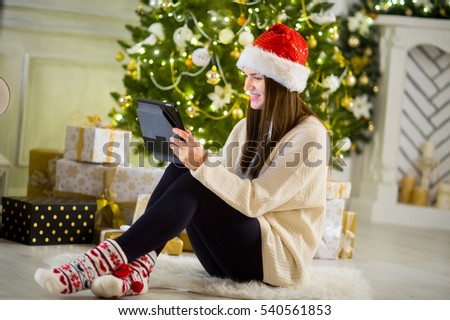 Charming girl in Santa's cap sits nearby a Christmas tree with the laptop. She attentively looks at the screen. Under an elegant Christmas tree there are a lot of beautiful boxes with gifts.