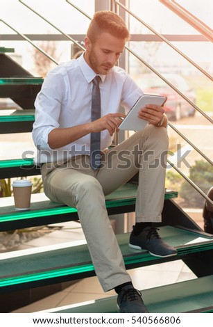 Young handsome businessman sitting on stairs with tablet and drinking coffee