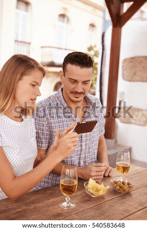 Blond girl showing young guy something on her cellphone with him looking at it with interest while out drinking some beer