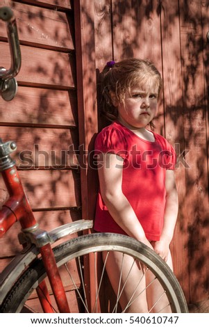 Cute little upset girl with vintage bicycle standing near the wooden wall of rustic house.