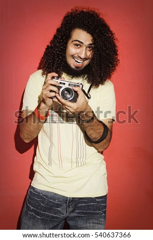Closeup of one handsome passionate expressive cool young brunette photographer men with long curly hair holding a vintage SLR camera standing against red background