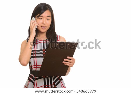 Young beautiful Asian businesswoman reading clipboard while talking on mobile phone isolated against white background