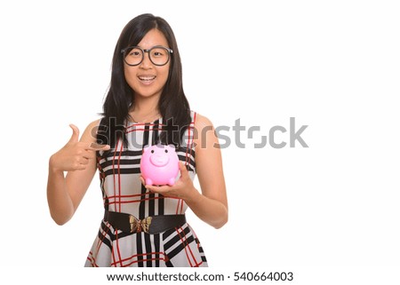 Young happy Asian businesswoman smiling and holding piggy bank and pointing finger isolated against white background
