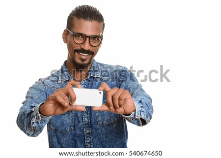 Young happy Indian man smiling and taking picture with mobile phone isolated against white background