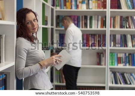 Beautiful young redhead girl reading a book in library. Doing research, thinking. Bookshelves with lots of books in background. 