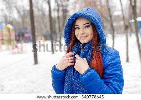 Pretty white brunette girl blow snow flakes from her frozen hands in snowy winter park.Good cold weather.Attractive young female wear knit earmuffs on her head,warm red scarf and white gloves
