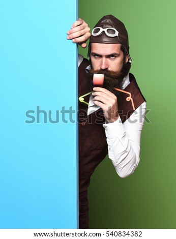 handsome bearded pilot or aviator man with long beard and mustache on serious face holding glass of alcoholic shot in vintage suede leather waistcoat with hat and glasses on colorful studio background
