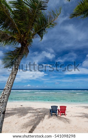 Coco Palm Garden Beach in Guam, USA. There are a pair of chairs (red and blue) on the white sand and a palm tree on the beach contiguous to the coral reef with transparent water.