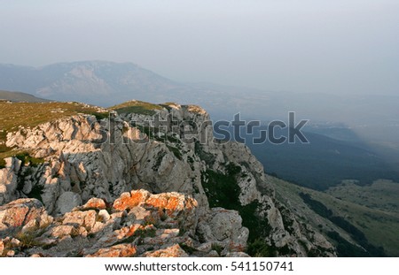 There are white limestone rocks with red lichens and distant mountains in gauze in the summer evening. This photo was taken in mountainous massif Chatyr-Dag, Crimea.