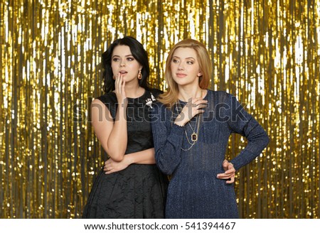 Two best friends having fun at New Year party. Young elegant women gossiping while standing over golden shining background