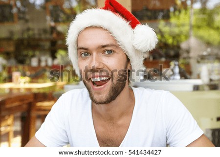 Close up shot of happy young hipster with stylish beard wearing white t-shirt and red Santa Claus hat, celebrating Christmas in hot tropical country, relaxing at cafe against blurred background