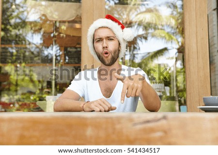 Headshot of stylish young man with stubble wearing Santa Claus hat having fun indoors, showing gesture with his finger and shouting greetings at camera, spending New Year's Day away in hot country