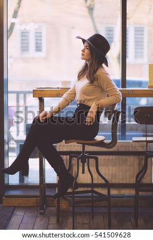 Attractive woman wearing in stylish wide-brimmed hat sitting on bar stool and drinking coffee in modern loft interior. Toned.