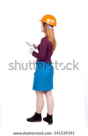 Backview of business woman in construction helmet stands and enjoys tablet or using mobile phone. Isolated over white background. Young woman engineer working on tablet. Engineer scorekeeper at work