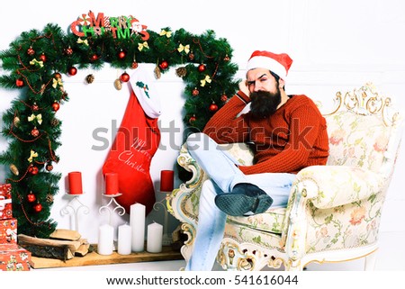 handsome bearded man with long beard in christmas red hat and sweater sitting bored on beige vintage armchair around christmas decoration on white studio background