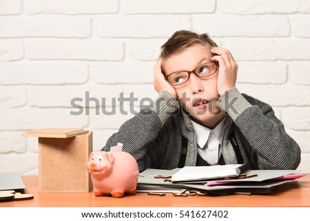 young cute pupil boy in grey sweater and glasses sitting at desk leaned on hands with papers pink piggy pig bank and wooden box in classroom on white brick wall background, copy space