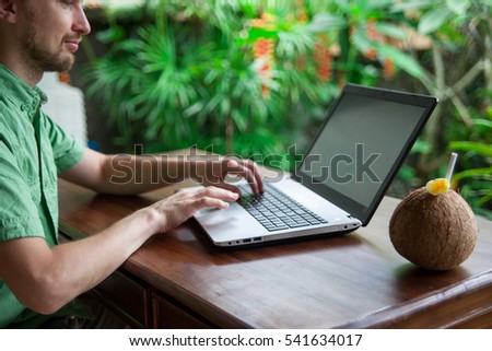 Handsome man freelancer typing on laptop on background of tropical garden. Distance work through internet concept. Soft focus background