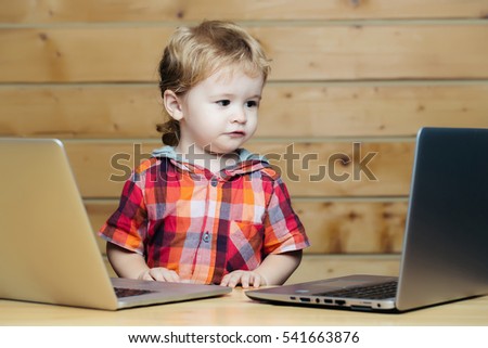 Cute baby boy child with blond curly hair plays on two laptop computers on wooden background