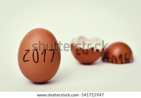 closeup of a whole brown chicken egg with the number 2017, as the new year, and a broken egg with the number 2016, as the old year, on an off-white background